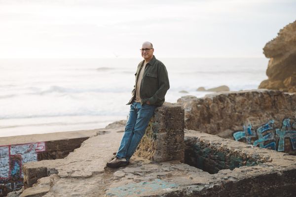 Balding man with glasses leaning up on old stone ruins in front of the Pacific Ocean.