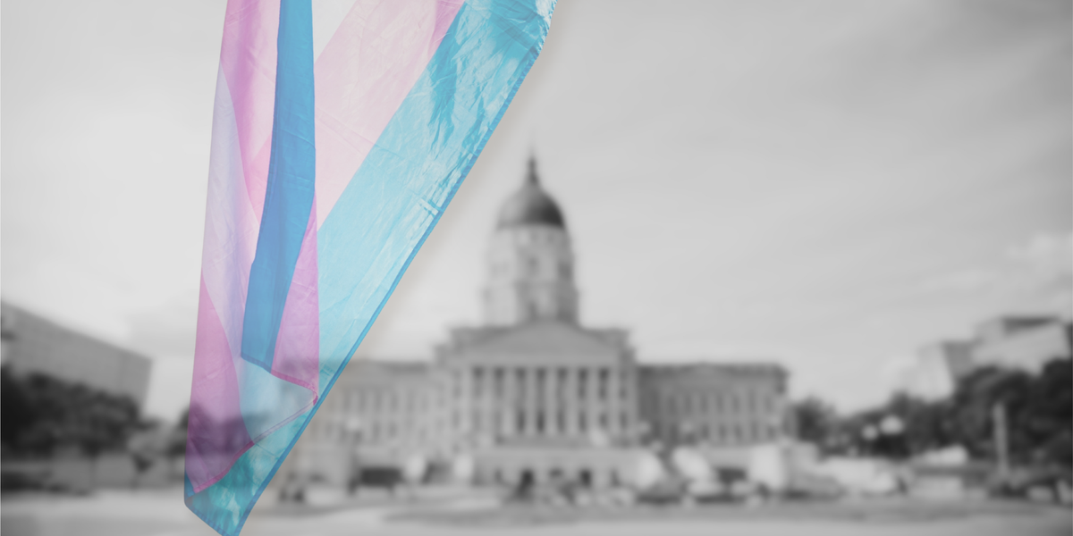 A Transgender Pride flag drapes mid-frame, backgrounded by a black and white photo of the Kansas State Legislature.