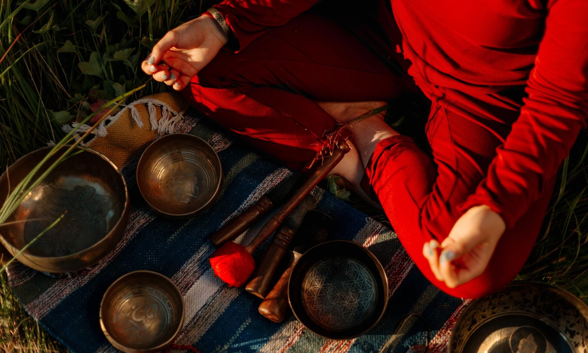 A photo of someone sat cross legged meditating outdoors, directed towards the ground where there are various bowls and tools.