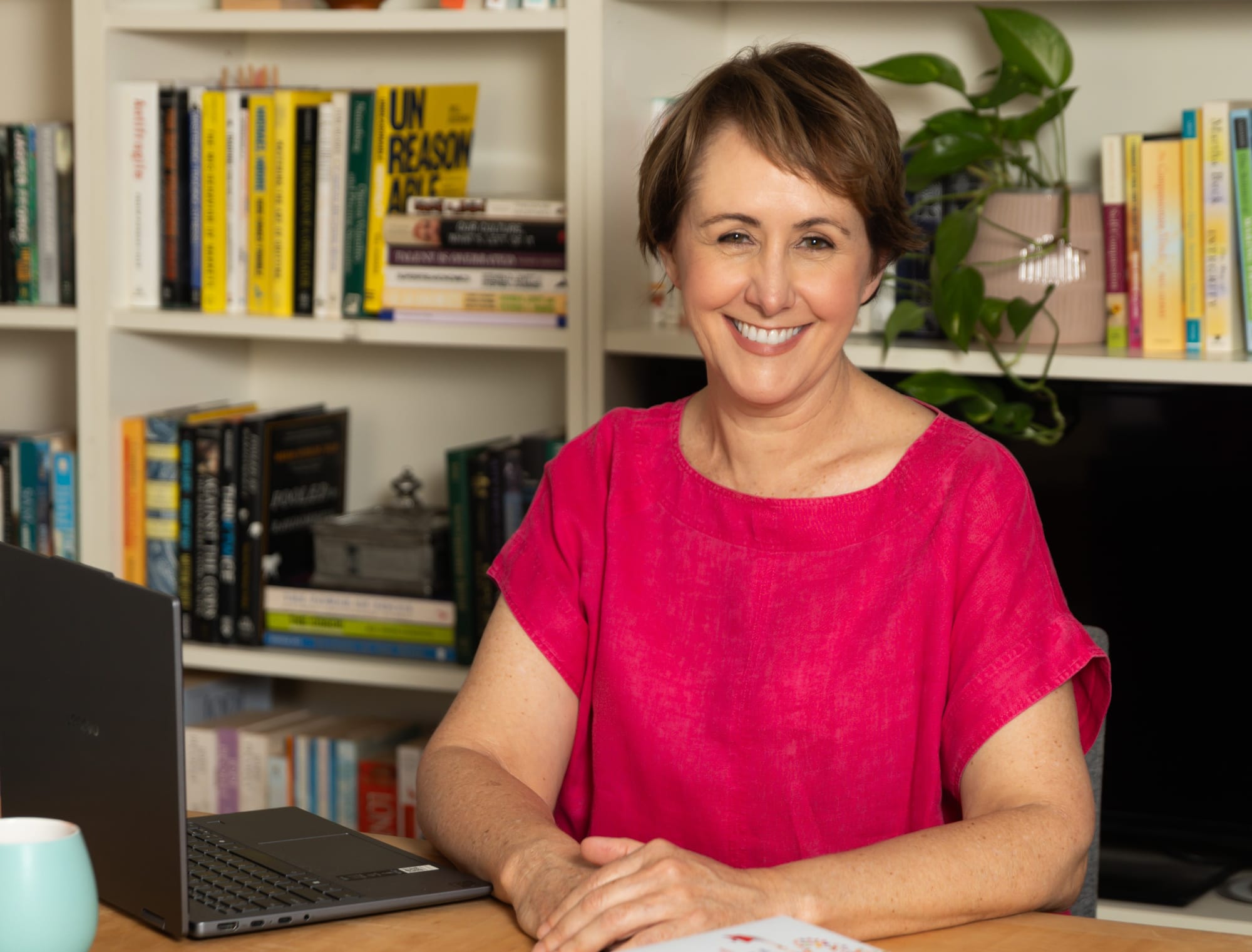 Photograph of a smiling woman sitting behind a desk with her hands folded in front of her, wearing a pink shirt. There is an open laptop in the foreground of the photo.