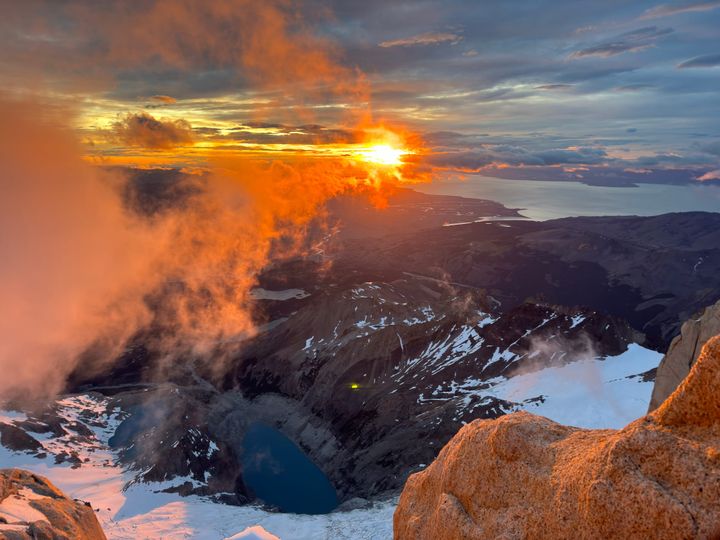 A cloudy sunrise over Laguna Sucia below Cerro Chaltén, with the town of El Chaltén and Lago Viedma in the distance.