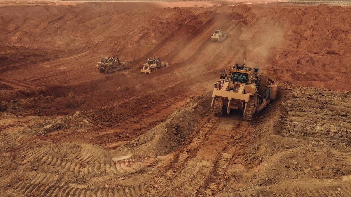 Vista aérea de quatro tratores amarelos em diferentes níveis de uma encosta de terra vermelha em mina brasileira. Máquinas levantam poeira, evidenciando a força bruta da extração mineral.