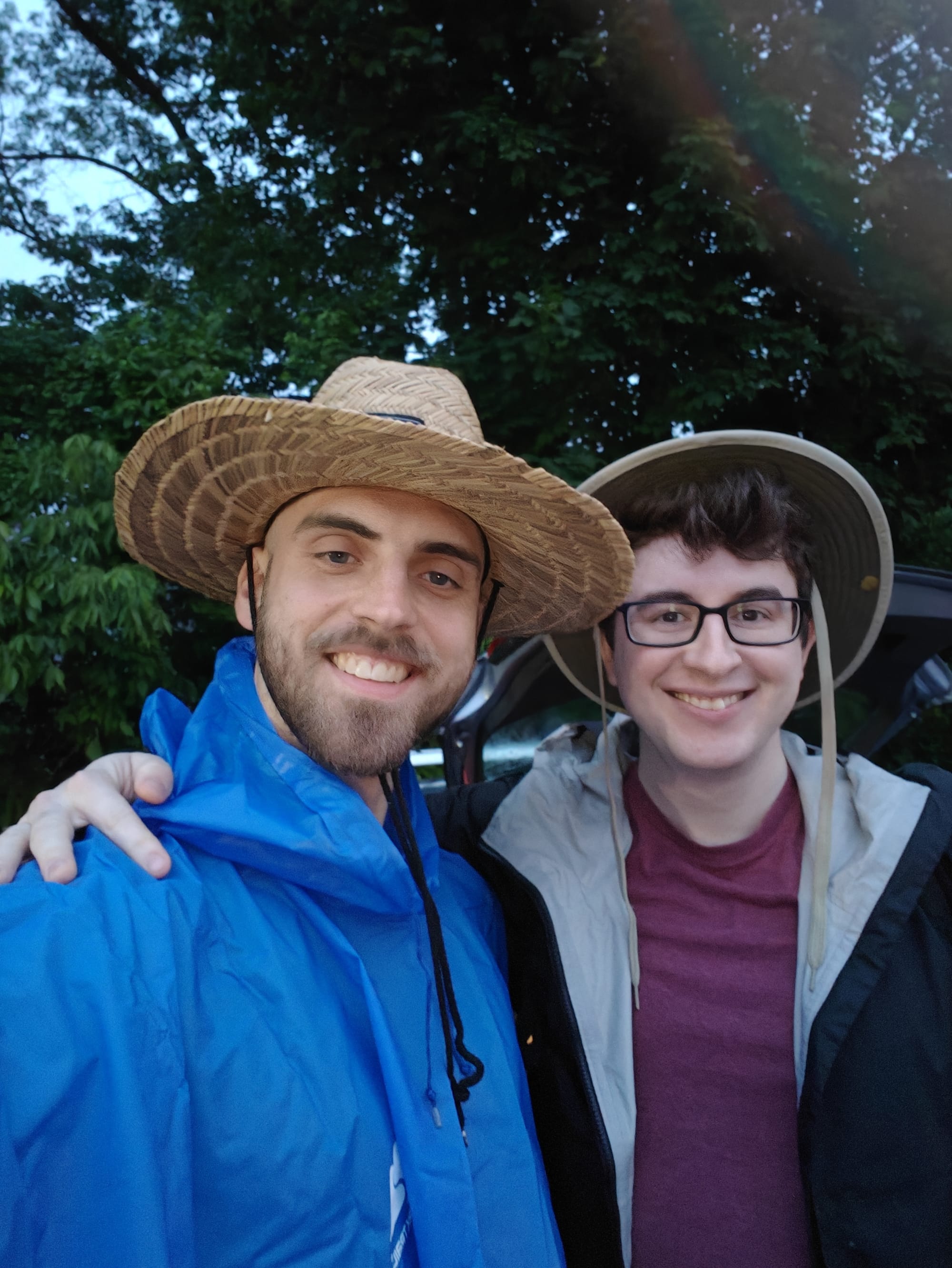 Two smiling men wearing wide-brimmed hats and jackets stand close together outdoors with trees in the background.