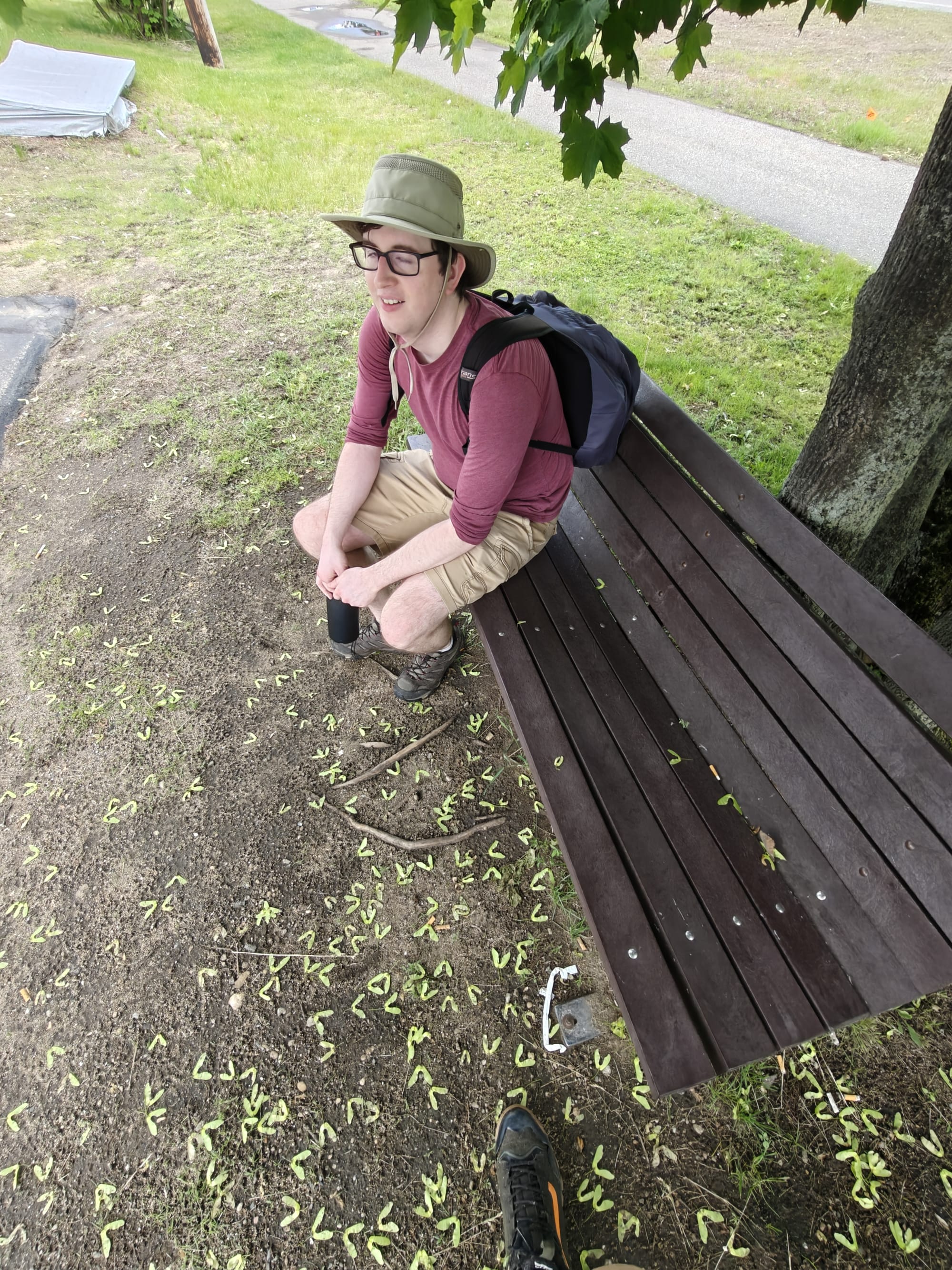 Alec sits on a bench wearing a hat, glasses, backpack, and casual clothes, with seeds scattered on the ground.