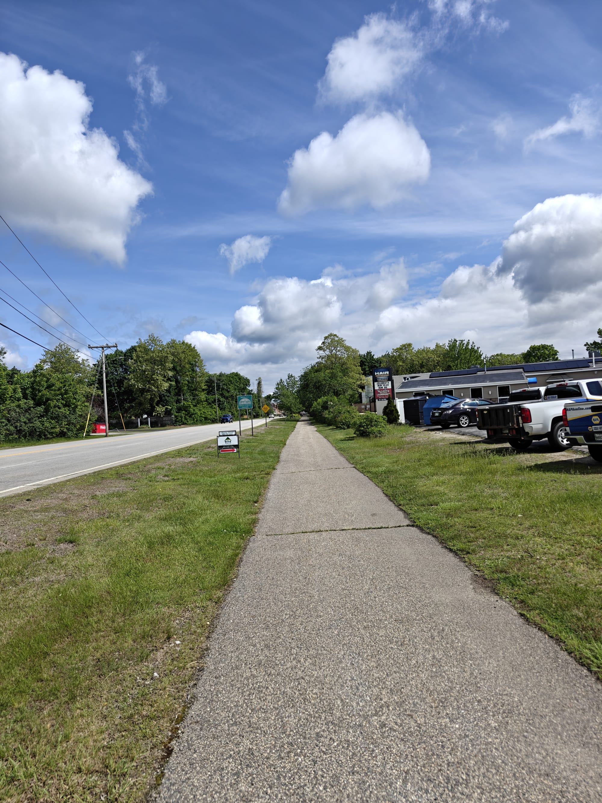 Paved sidewalk alongside a road with trees and commercial buildings under a blue sky with clouds.