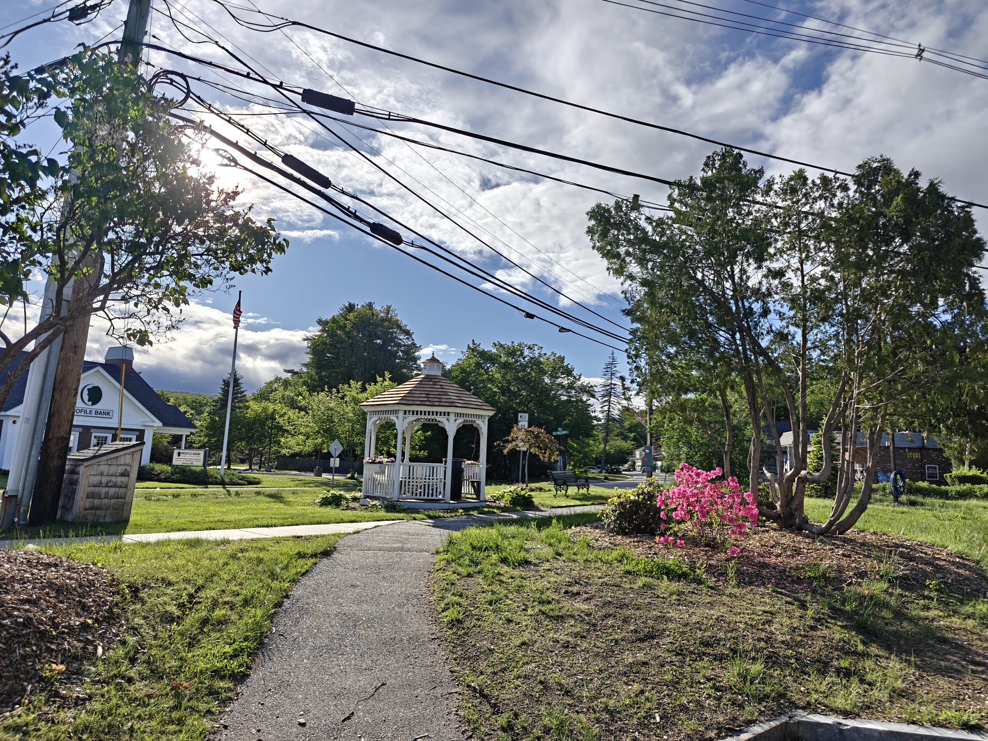 Scenic village common area with a white gazebo, sidewalk, trees, and blooming pink flowers.