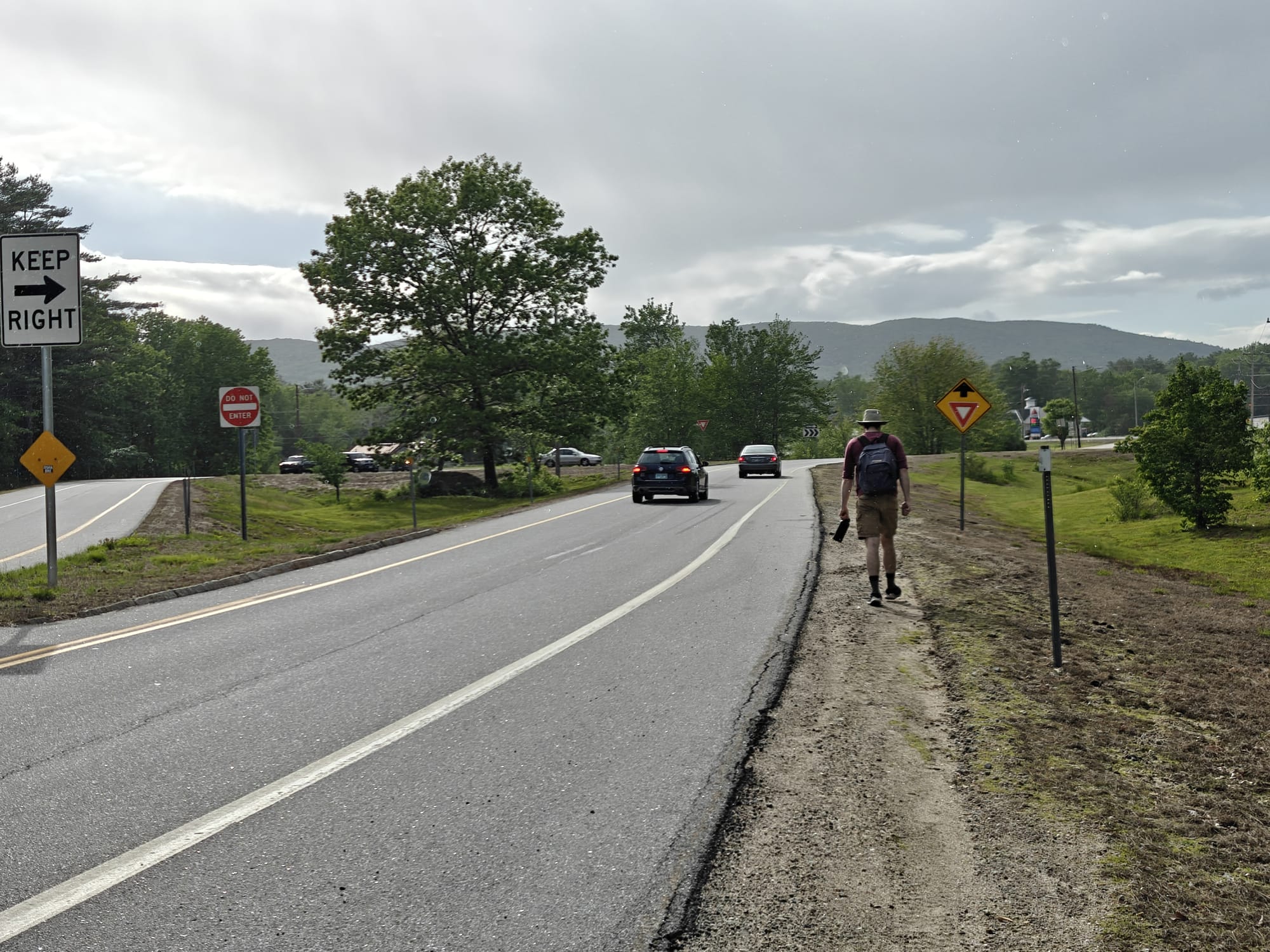 Alec walks on grassy road shoulder beside traffic, signs, trees, and distant mountains.