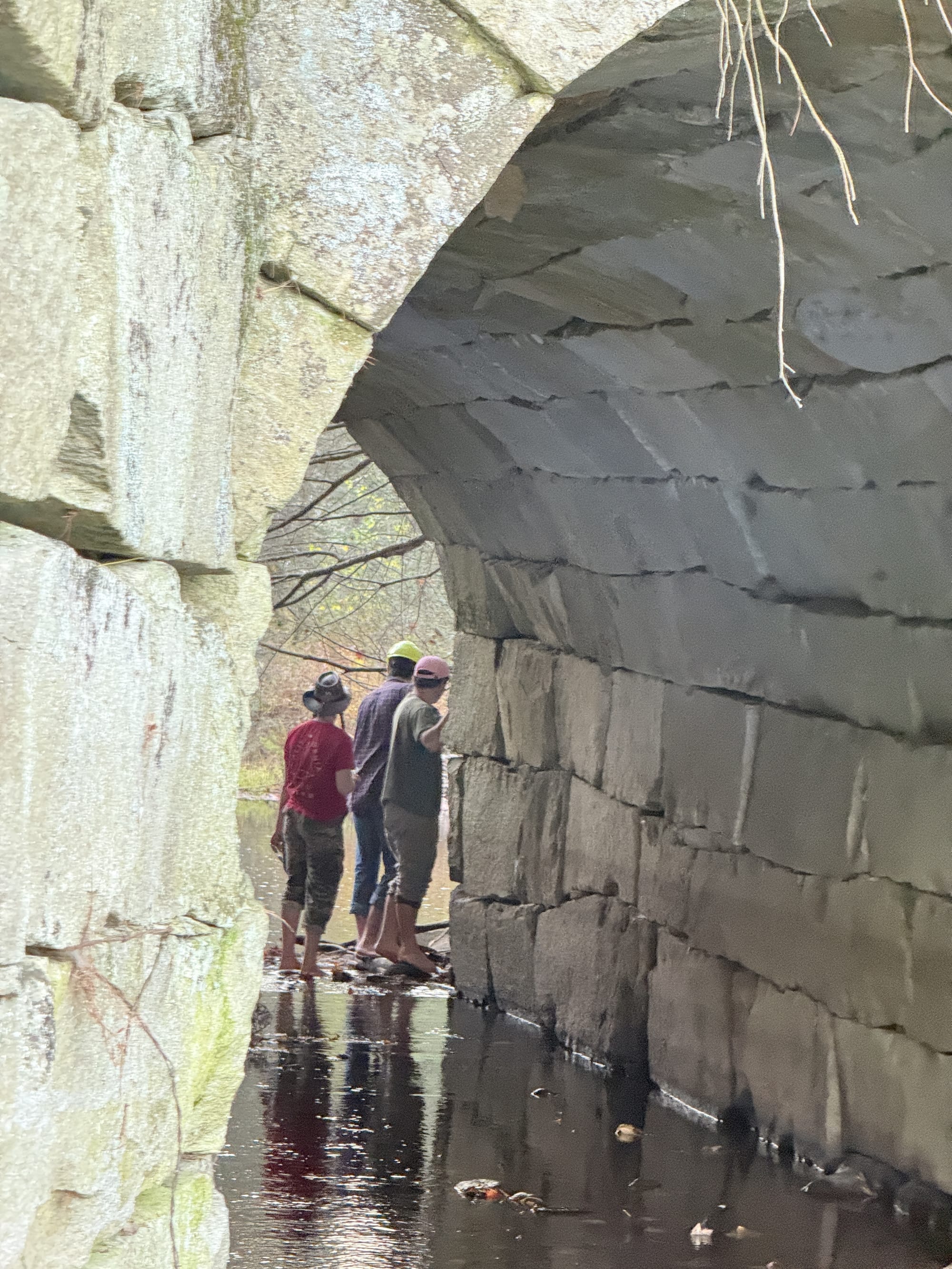 Three friends, including one in a hardhat, stand barefoot in shallow water under an old stone railroad tunnel over Blackwater Brook.