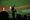 A spectator in silhouette leans over a glass railing and raises an arm toward a baseball field during a night game, with two players standing on the infield below.