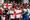 A jubilant crowd of England football fans on sunny stone steps, arms raised and cheering, waving St George's Cross flags.