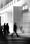 Two people in business attire talk outside a glass office building as a blurred figure walks past.