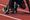 A sprinter crouched in the starting blocks on a red running track, one hand holding a relay baton behind the white start line, poised to push off.