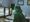 A masked clinician in green scrubs and goggles works at a hospital workstation lined with monitors showing medical scans.