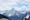 A snow-covered mountain peak rising above a rocky ridge under a partly cloudy sky, with a small stack of stones in the foreground.