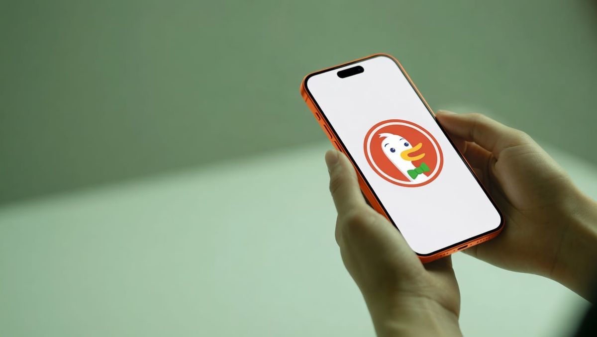 Hands holding an orange iPhone with the DuckDuckGo mascot Dax the Duck on screen against a green background