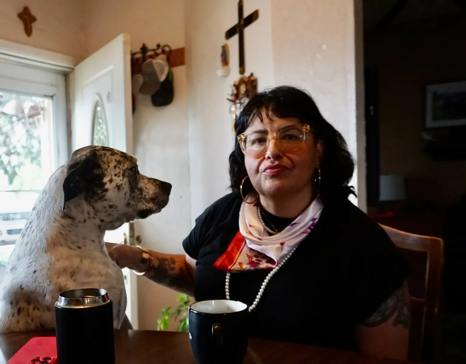 Woman wearing glasses sits at a wooden table with a black coffee mug, petting a white and black-spotted dog inside a home.
