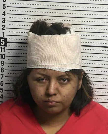 Booking photo of a woman wearing a red jail uniform and a white bandage wrapped around her head, standing in front of a height chart.