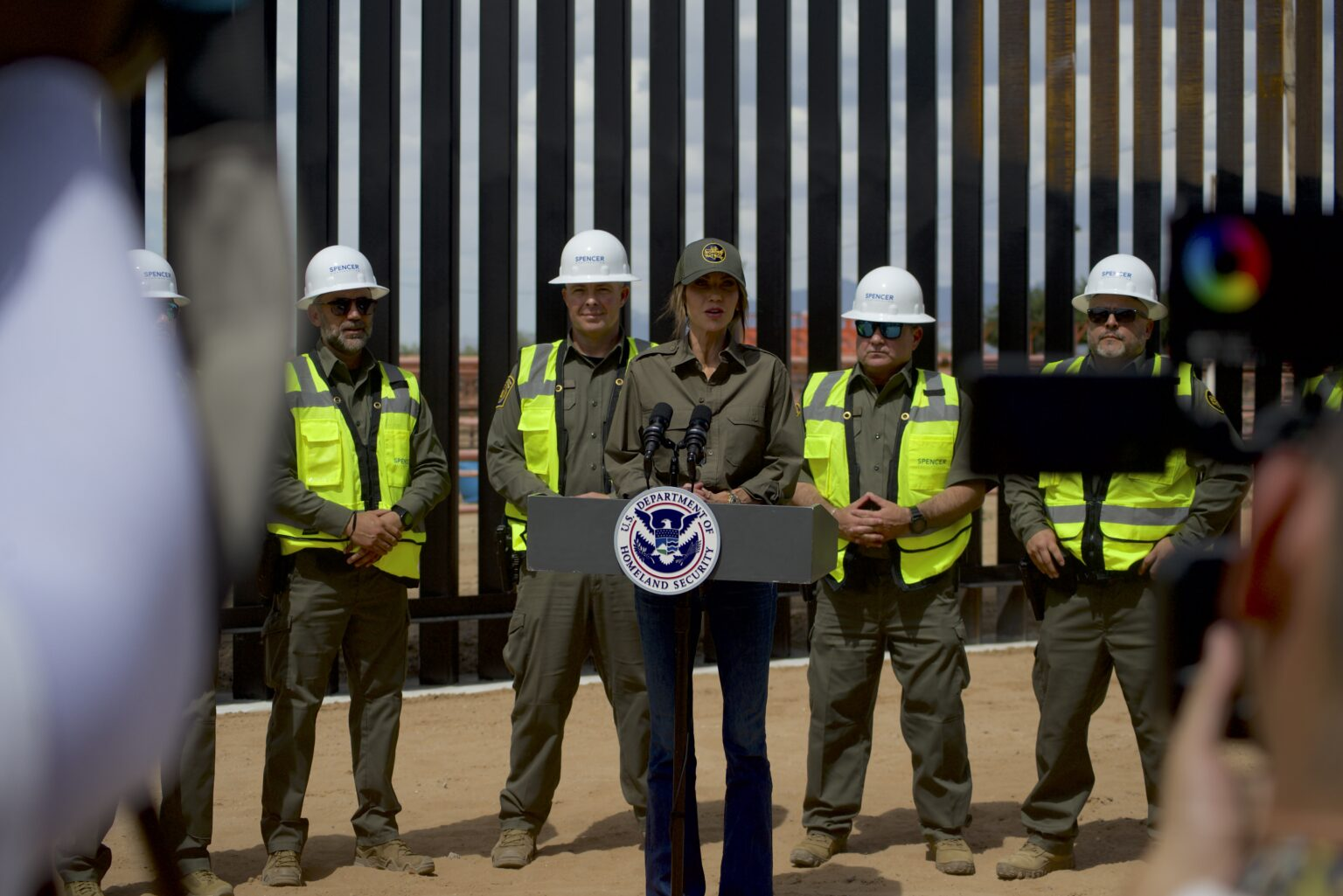 Homeland Security Secretary Kristi Noem speaks at a podium in front of the border wall in Santa Teresa, flanked by uniformed officials and workers in safety vests and hard hats.