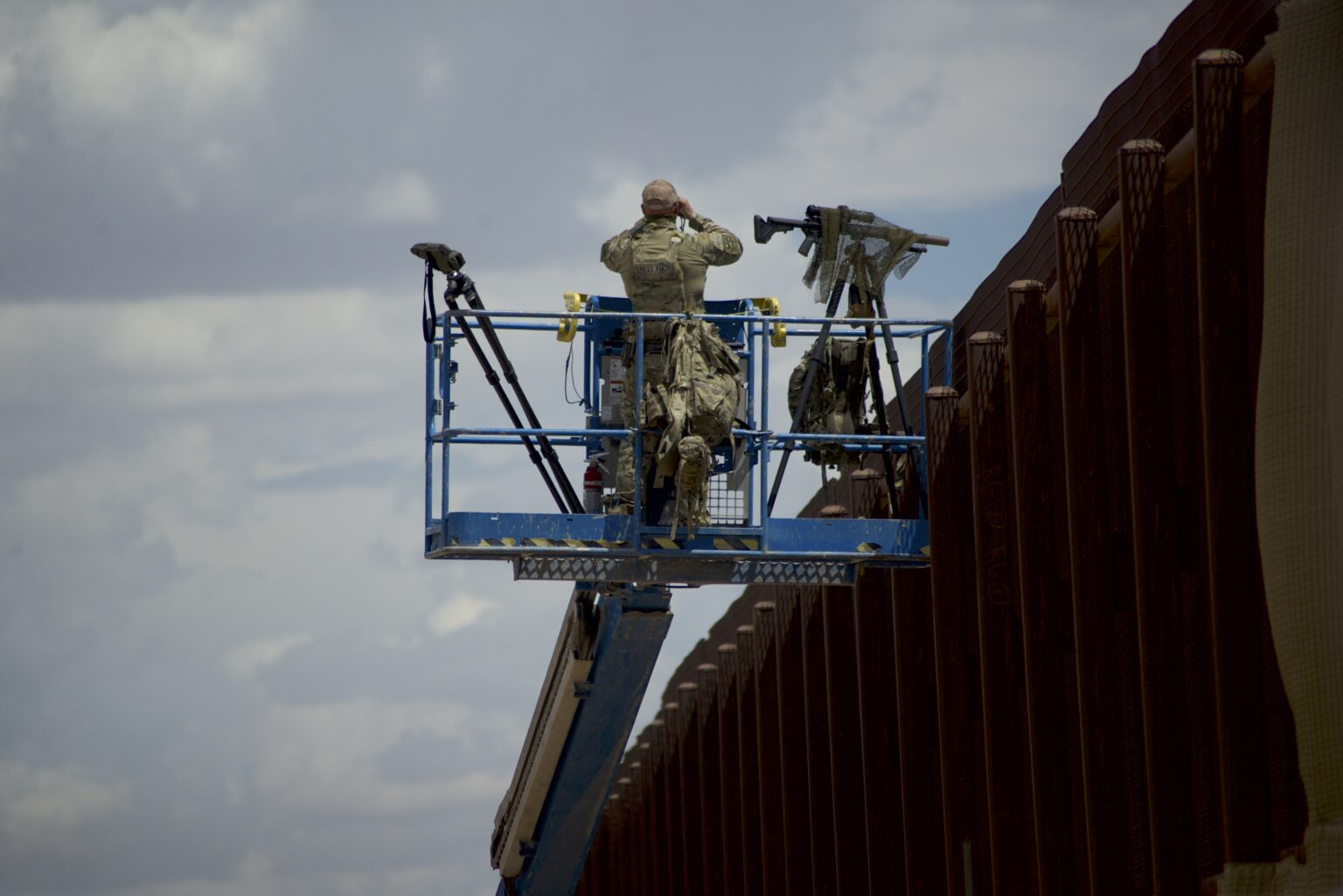 Border Patrol agent stands on a blue lift platform alongside equipment, scanning the U.S.-Mexico border wall with binoculars.