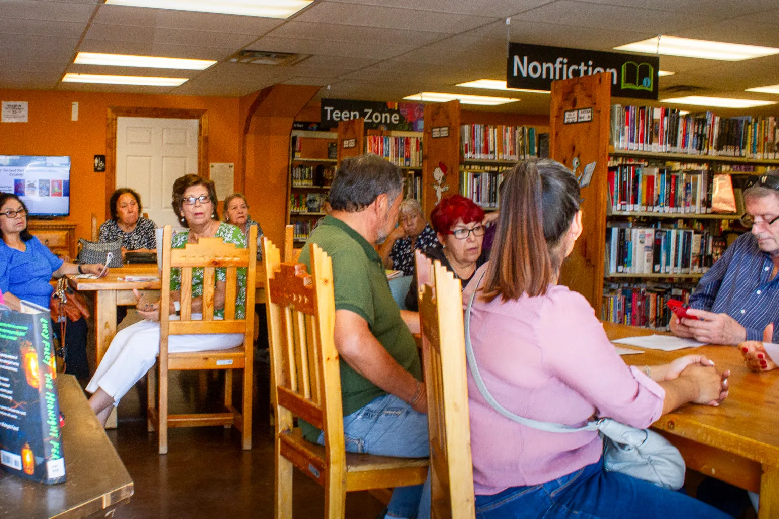 Community members gather for a discussion inside a library, seated at wooden tables beneath signs for the Teen Zone and Nonfiction sections.
