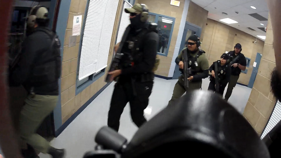Armed detention officers in tactical gear move in formation down a hallway inside the Doña Ana County Detention Center during a training or raid scenario.