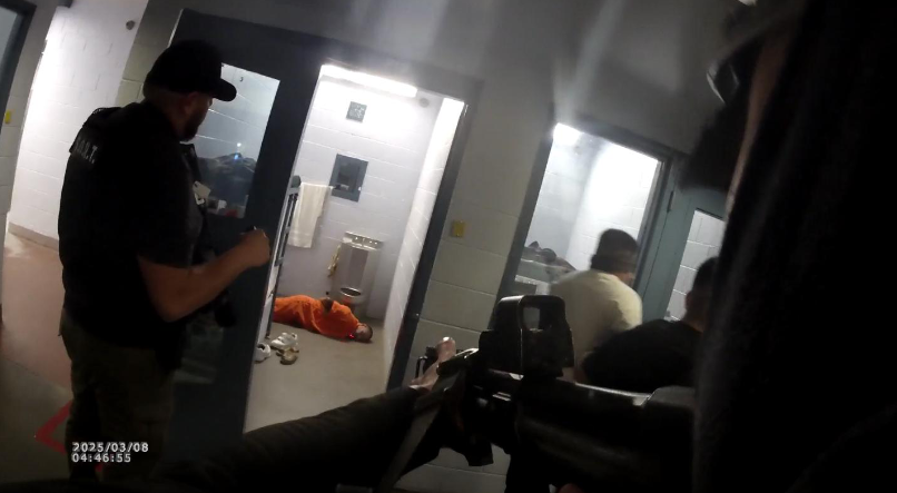 Detention officers armed with rifles stand outside a cell where a man in an orange jumpsuit lies face down on the floor at the Doña Ana County Detention Center during a raid or training scenario.