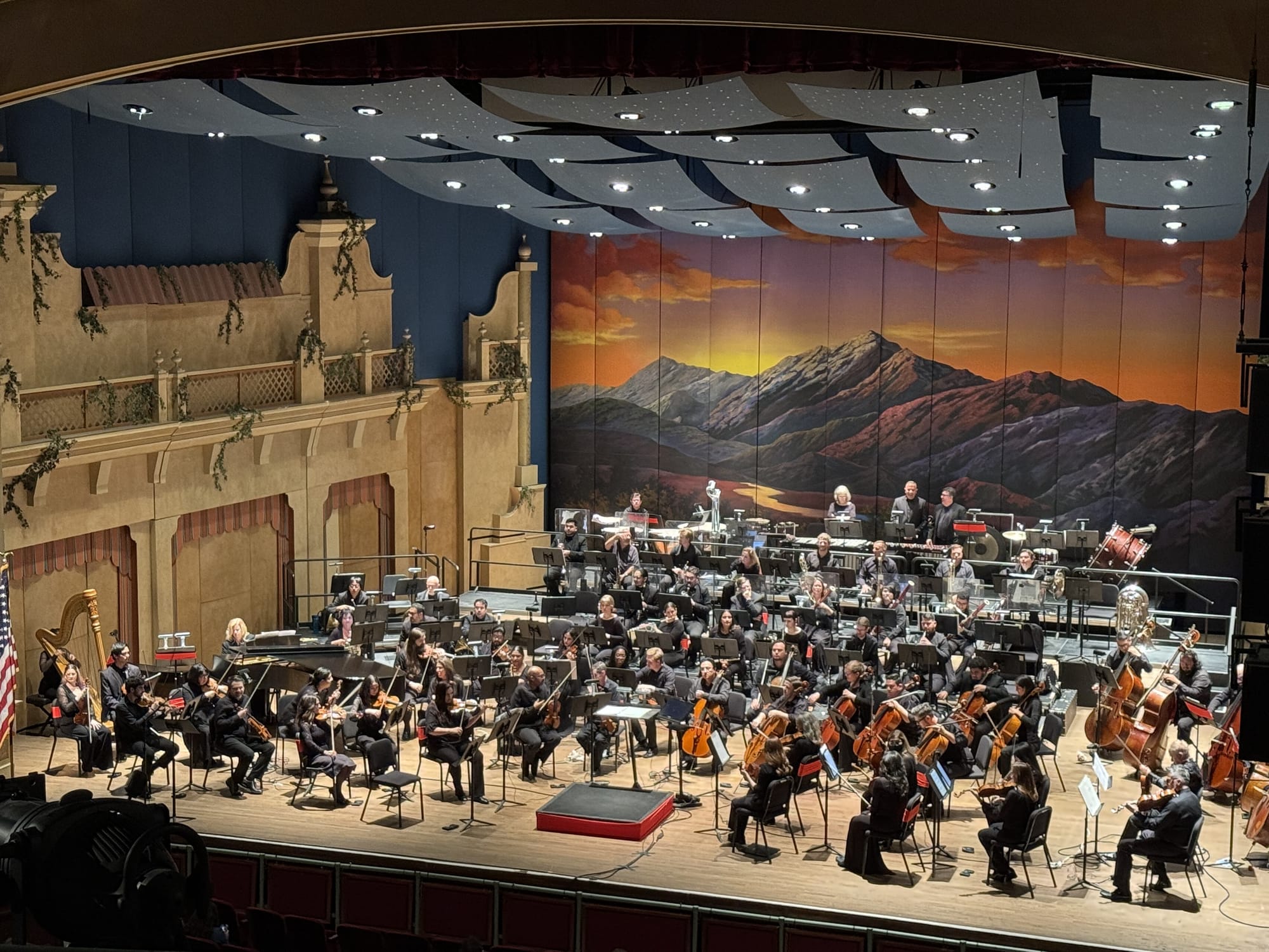 The El Paso Symphony Orchestra performs onstage at the Plaza Theatre, with musicians arranged in sections beneath a backdrop depicting mountains at sunset.