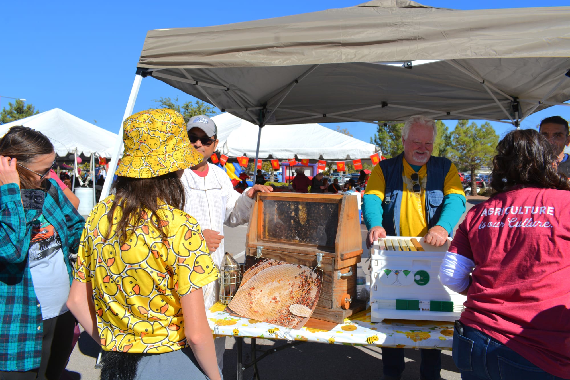 A man shows a group of people a live honeybee display under a canopy during Ag Day at New Mexico State University, with colorful tents and booths in the background.
