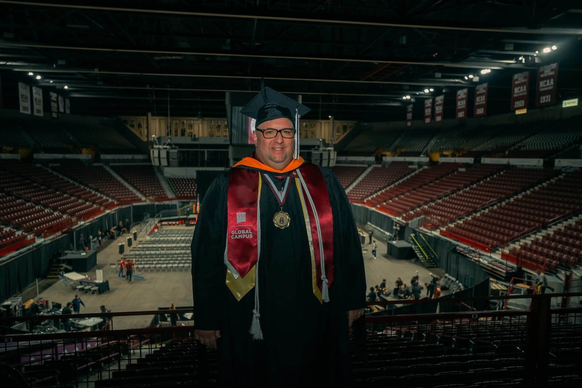A graduate wearing an NMSU Global Campus stole stands inside the Pan American Center in Las Cruces following commencement, with the arena’s stage and seating visible in the background.