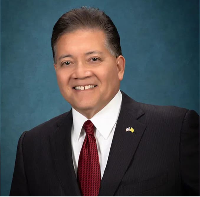 Former Las Cruces mayor Ken Miyagishima in a dark suit and red tie smiling in front of a blue background.