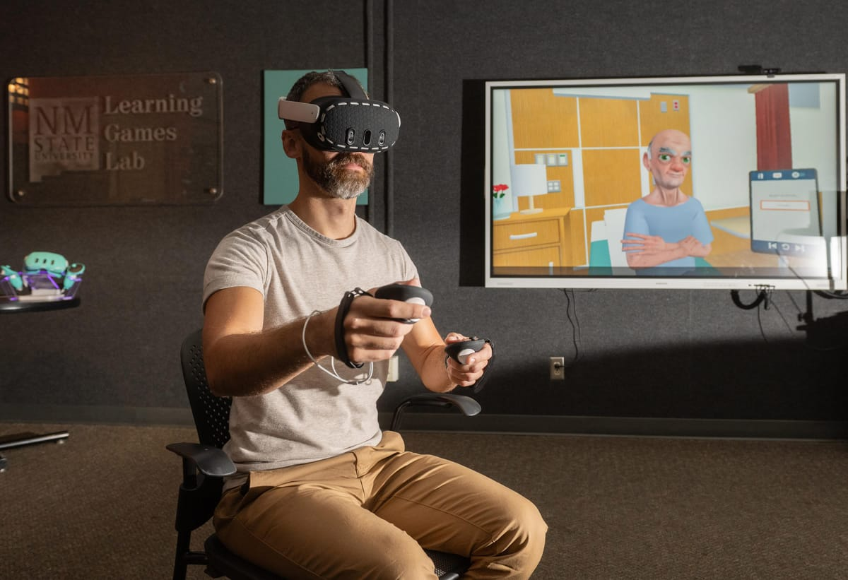 A man wearing a virtual reality headset and hand controllers participates in a nutrition-training simulation at NMSU’s Learning Games Lab, with an animated patient shown on a nearby screen.