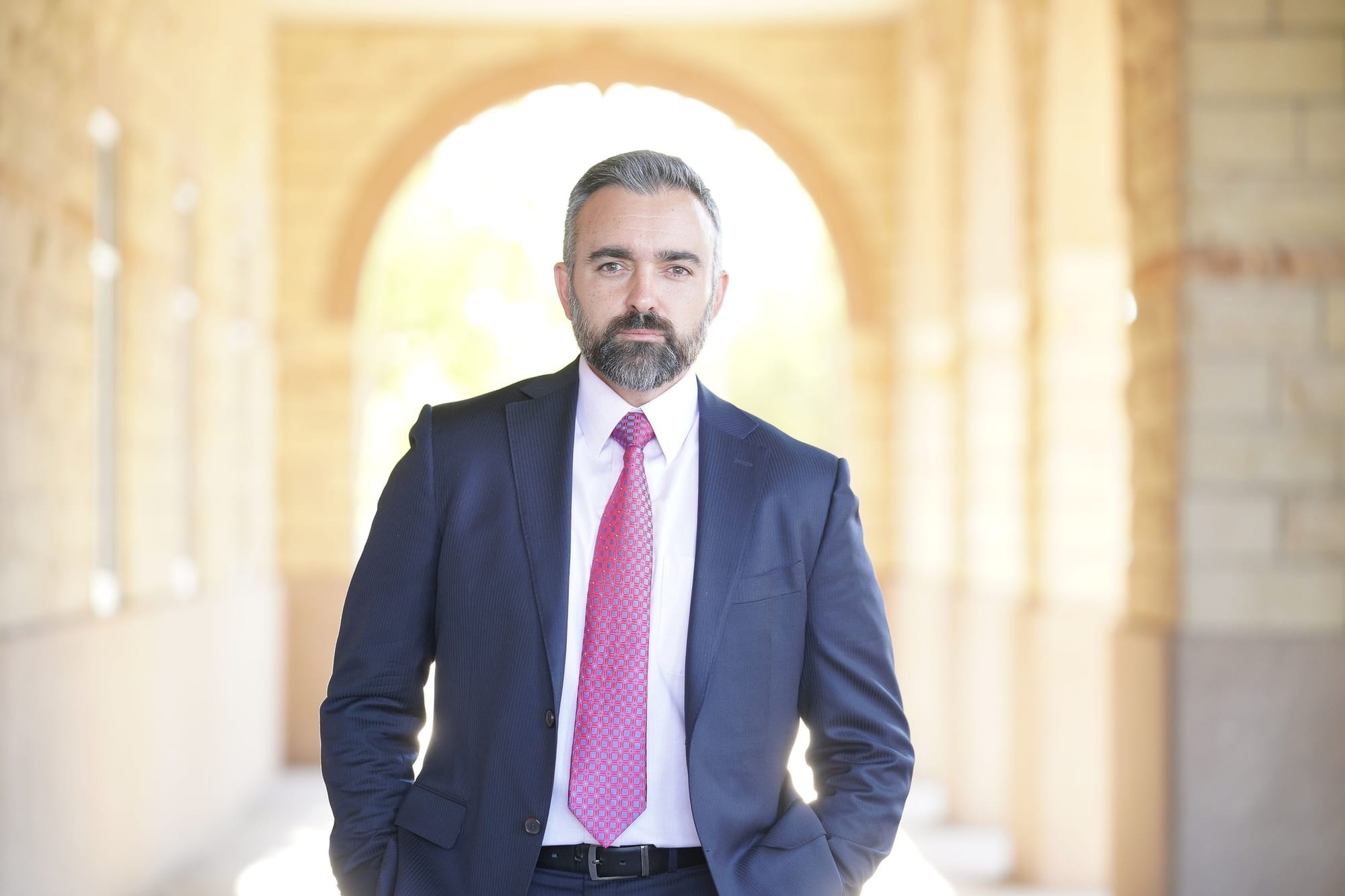 Attorney General Raúl Torrez, in a navy suit, white shirt and pink tie stands facing the camera inside a softly lit arched corridor with a bright background.