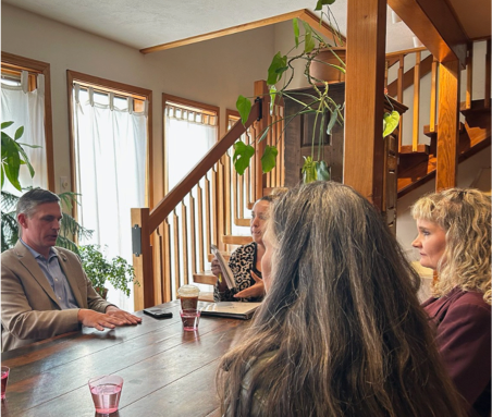 U.S. Senator Martin Heinrich sits at a dining table with three women in a sunlit home interior, discussing health care concerns during a small roundtable meeting.
