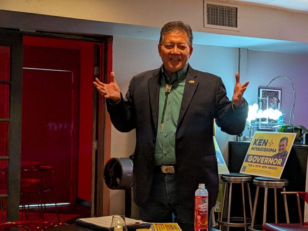 Former Las Cruces Mayor Ken Miyagishima speaks to supporters indoors, standing with his arms raised near campaign signs during his run for New Mexico governor.