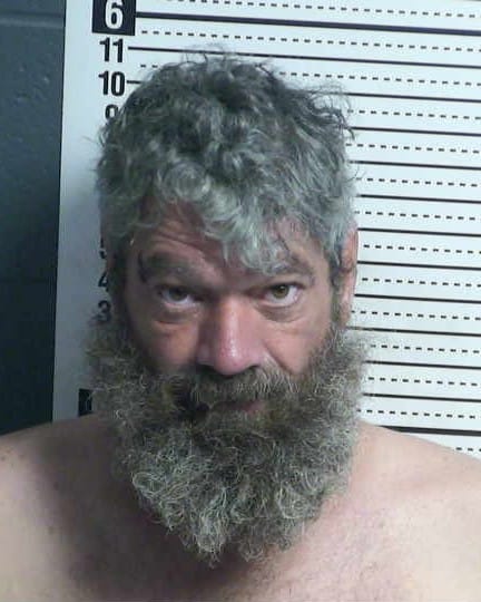 A booking photo of Brian Smolak, a middle-aged man with gray curly hair and a long beard, standing shirtless in front of a height chart at the Doña Ana County Detention Center.