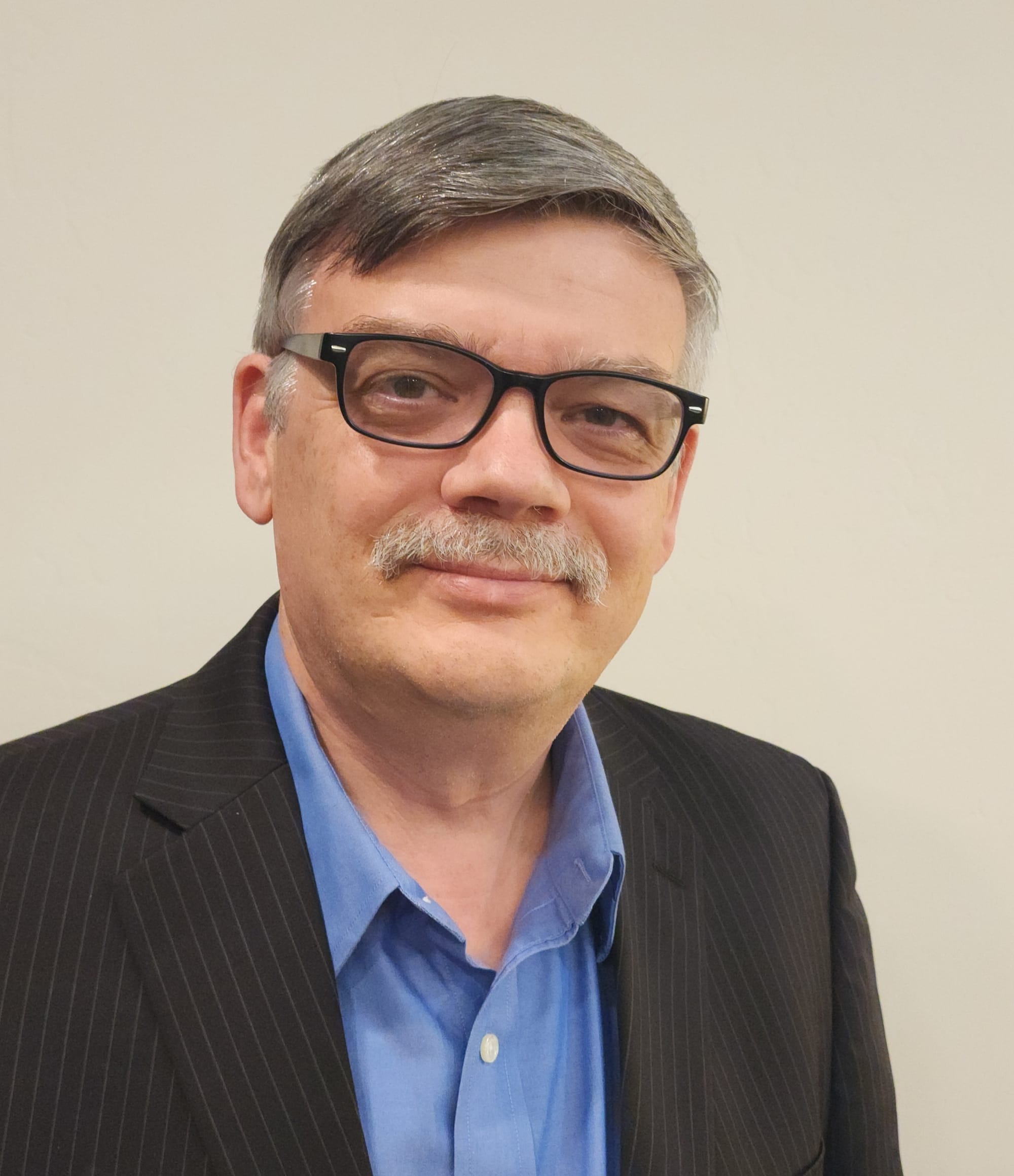 Head-and-shoulders portrait of Sergey Dryga wearing black-framed glasses, a blue collared shirt and a dark pinstriped blazer against a plain light background.