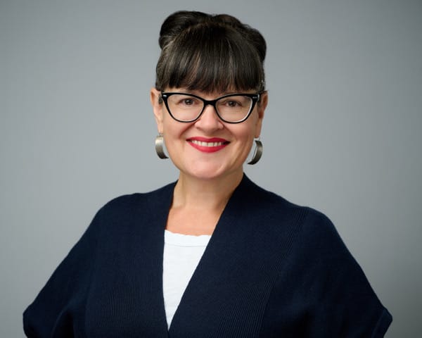 Gwyneth Doland, with dark hair in an updo, wearing glasses, silver earrings and a dark cardigan over a white shirt, smiles at the camera against a gray background.