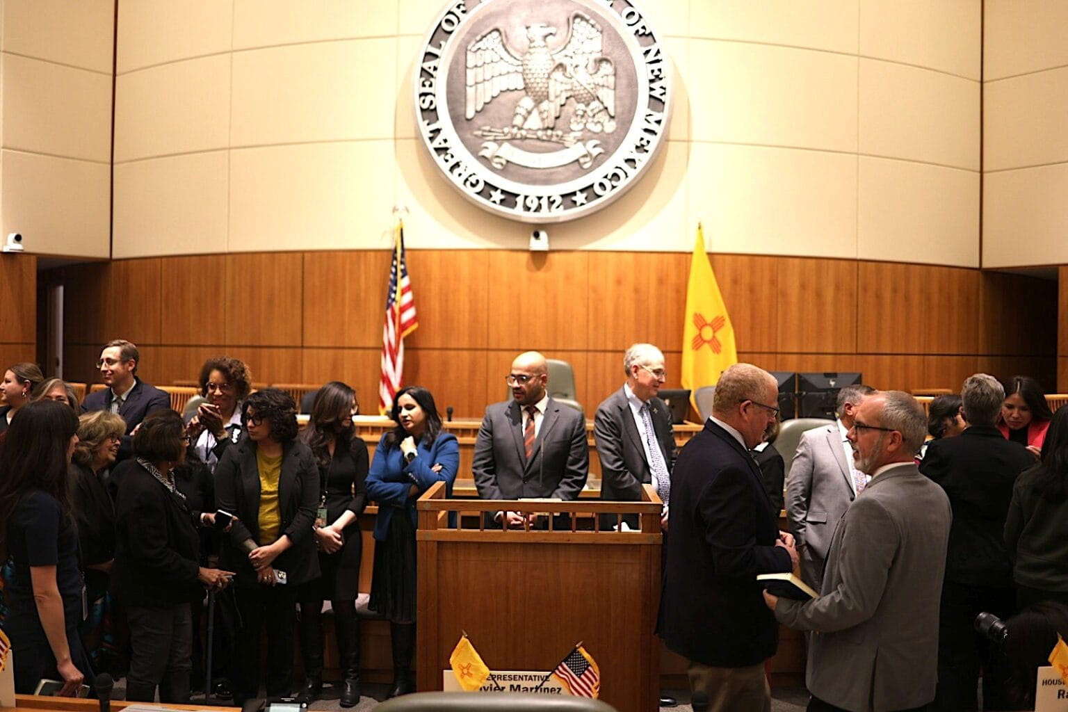 People gather and talk on the New Mexico House floor beneath the state seal and flags after the Nov. 10, 2025 special legislative session.