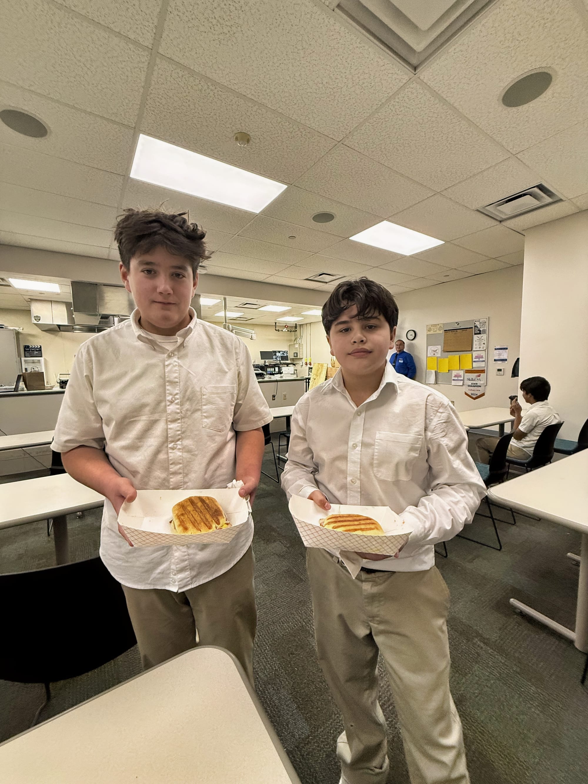 Two middle school students stand in a classroom holding trays with grilled Cubano paninis they prepared during a culinary arts visit at DACC.