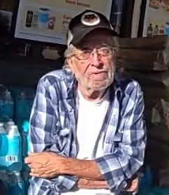 An older man with gray hair and glasses wears a black baseball cap, blue plaid shirt and white T-shirt while sitting indoors with his arms crossed. He uses a cane to help walk.