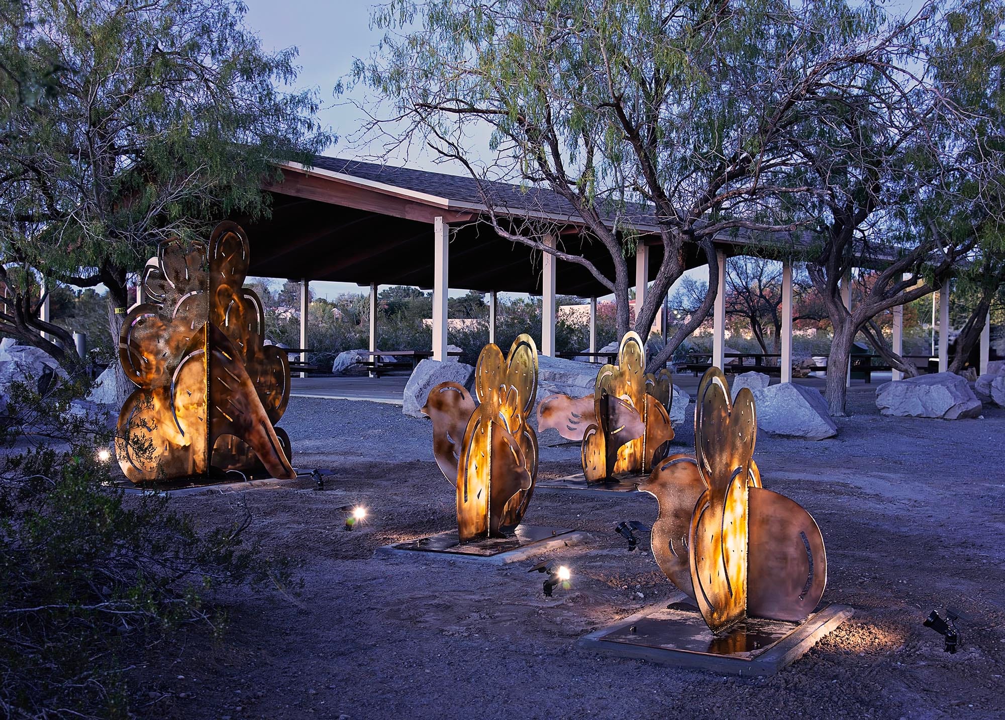 Illuminated metal sculptures inspired by desert plants are installed along a pathway at a Las Cruces park, with a covered picnic pavilion, trees and rocks visible at dusk.