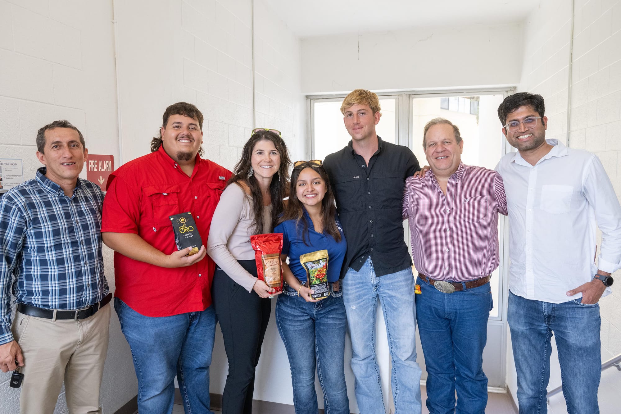 A group of seven people, including NMSU students and faculty, stand together smiling and holding bags of Puerto Rican coffee during a campus visit after their research trip.