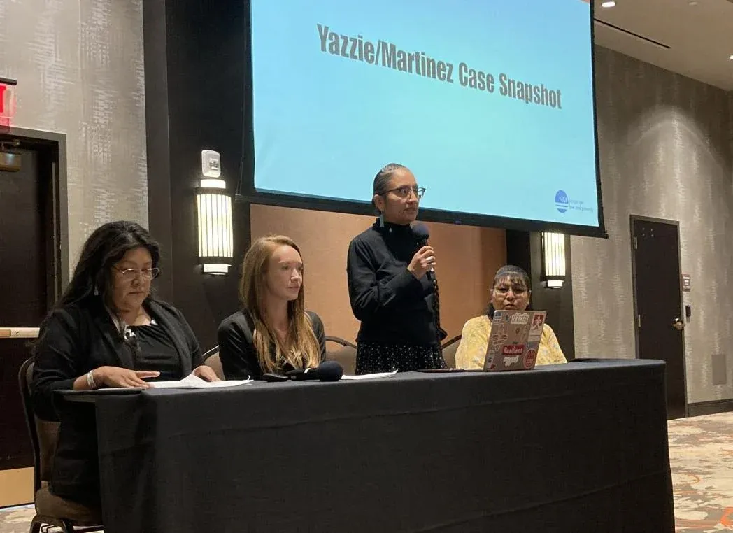 Four speakers sit at a panel table as a presenter stands with a microphone in front of a screen titled Yazzie/Martínez Case Snapshot during a public meeting.