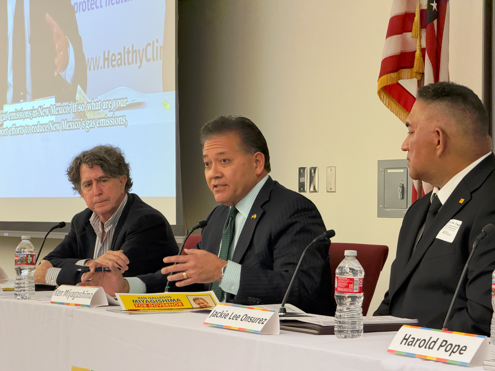 Ken Miyagishima speaks during a Healthy Climate New Mexico town hall panel, seated between other panelists at a table with microphones and name placards visible.
