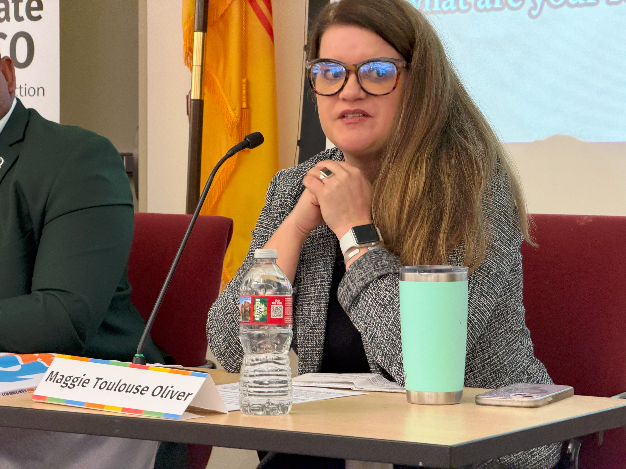 Maggie Toulouse Oliver speaks during a Healthy Climate New Mexico town hall panel, seated at a table with a microphone, name placard and notes visible in front of her.