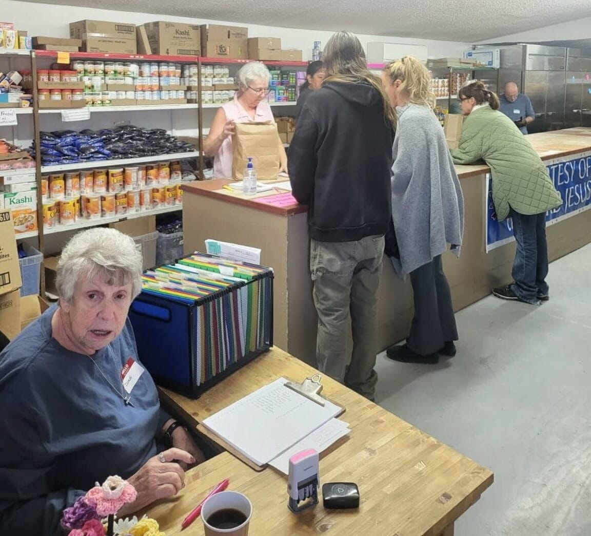 Volunteers at a food pantry prepare bags of groceries for people waiting at the counter, with shelves of canned goods and staples visible behind them.