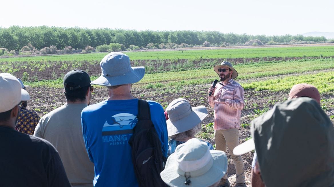 A man with a microphone speaks to a small group standing in a farm field, with rows of crops and trees visible in the background.