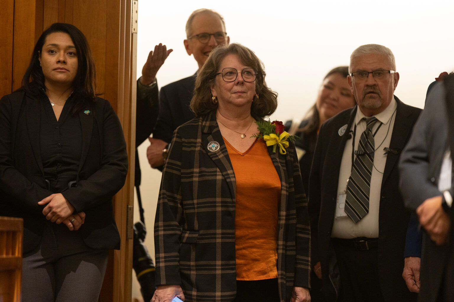 State Sen. Linda Trujillo stands indoors with other officials in a legislative building hallway, wearing an orange top and plaid jacket, as people gather behind her.