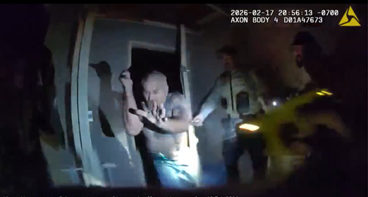 Still image from an Axon body camera showing deputies inside a darkened doorway as a man raises his arm while officers illuminate him with flashlights during a nighttime response.