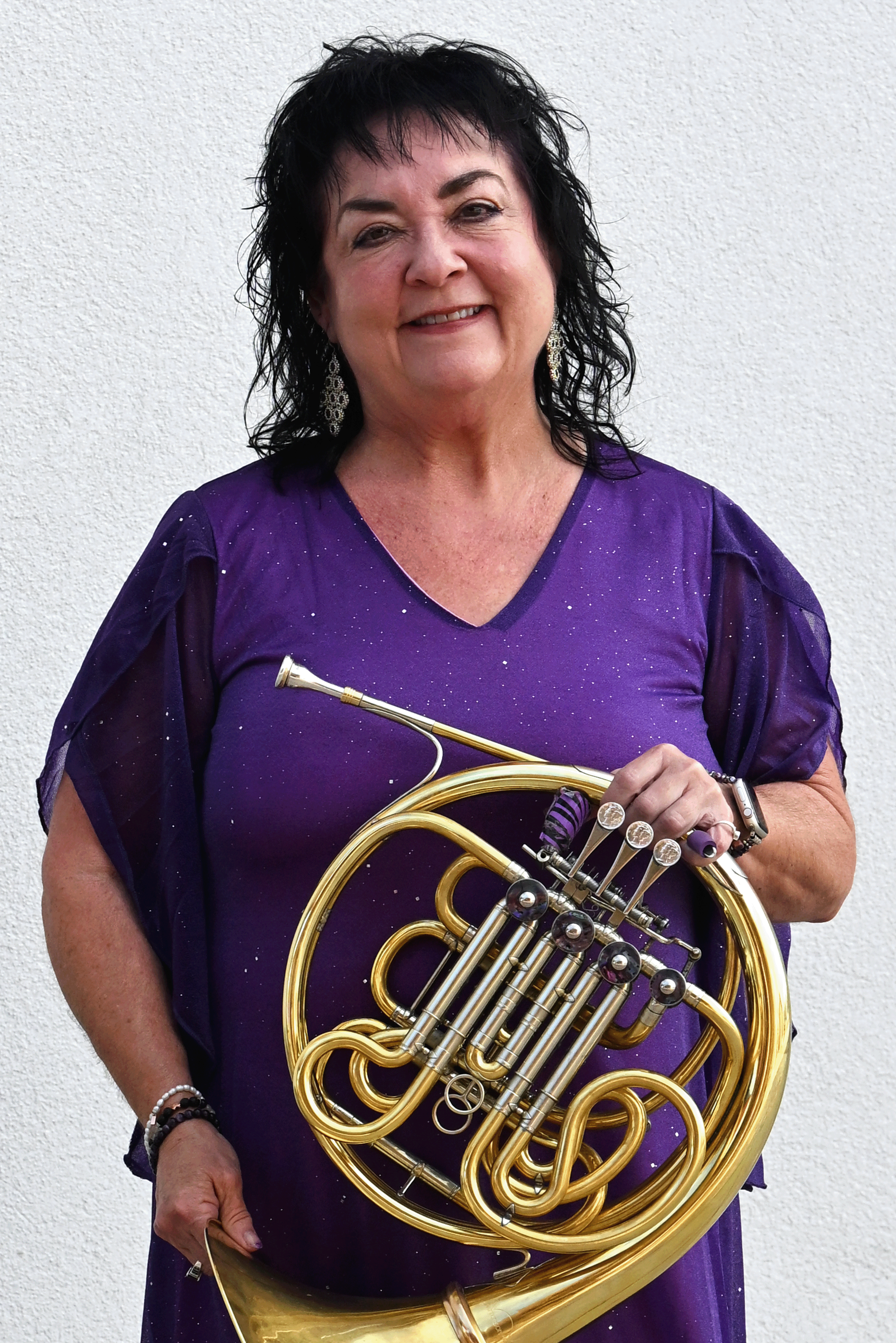 Nancy Joy, a French horn player and retired New Mexico State University horn professor, stands smiling against a light wall while holding a brass French horn and wearing a purple top.